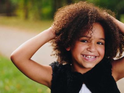 young girl with curly brown hair and hands on her head smiling