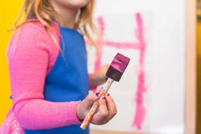 young girl wearing blue apron holding a paint brush