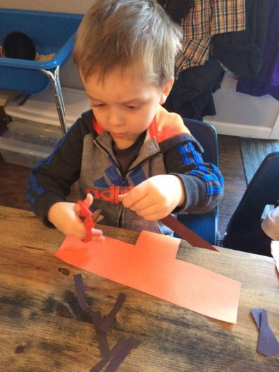 young boy cutting a piece of orange paper at the art table